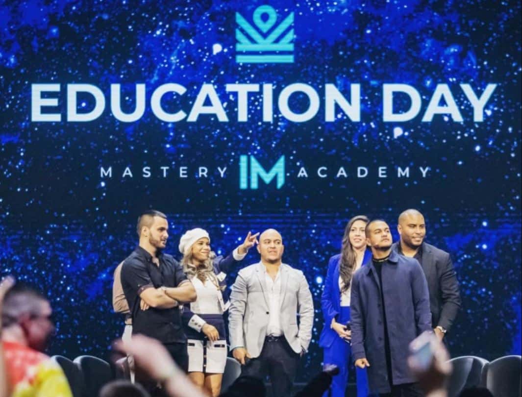 IM Mastery Academy (iMarketsLive) Erfahrungen – Chance oder Betrug? Mehrere Personen auf einer Bühne während eines 'Education Day' Events der IM Mastery Academy. Im Hintergrund eine große LED-Wand mit dem Schriftzug 'Education Day' und dem Logo der IM Academy. Die Bildunterschrift weist auf die Rekrutierung von Vertriebspartnern und den MLM-Charakter des Unternehmens hin.